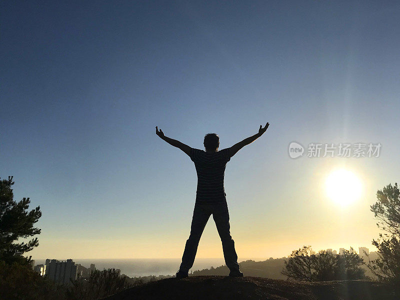在智利瓦尔帕莱索的维纳(Vina)，一名男子伸开双臂在山顶上庆祝，身后是太平洋的景色和日落，Viña del Mar, Reñaca